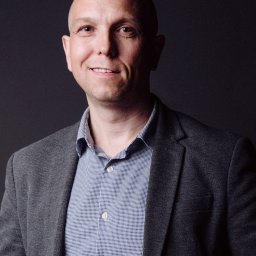 Sellflow Sp. z o.o. - Portret uśmiechniętego mężczyzny w szarej marynarce i niebieskiej koszuli na ciemnoszarym tle.