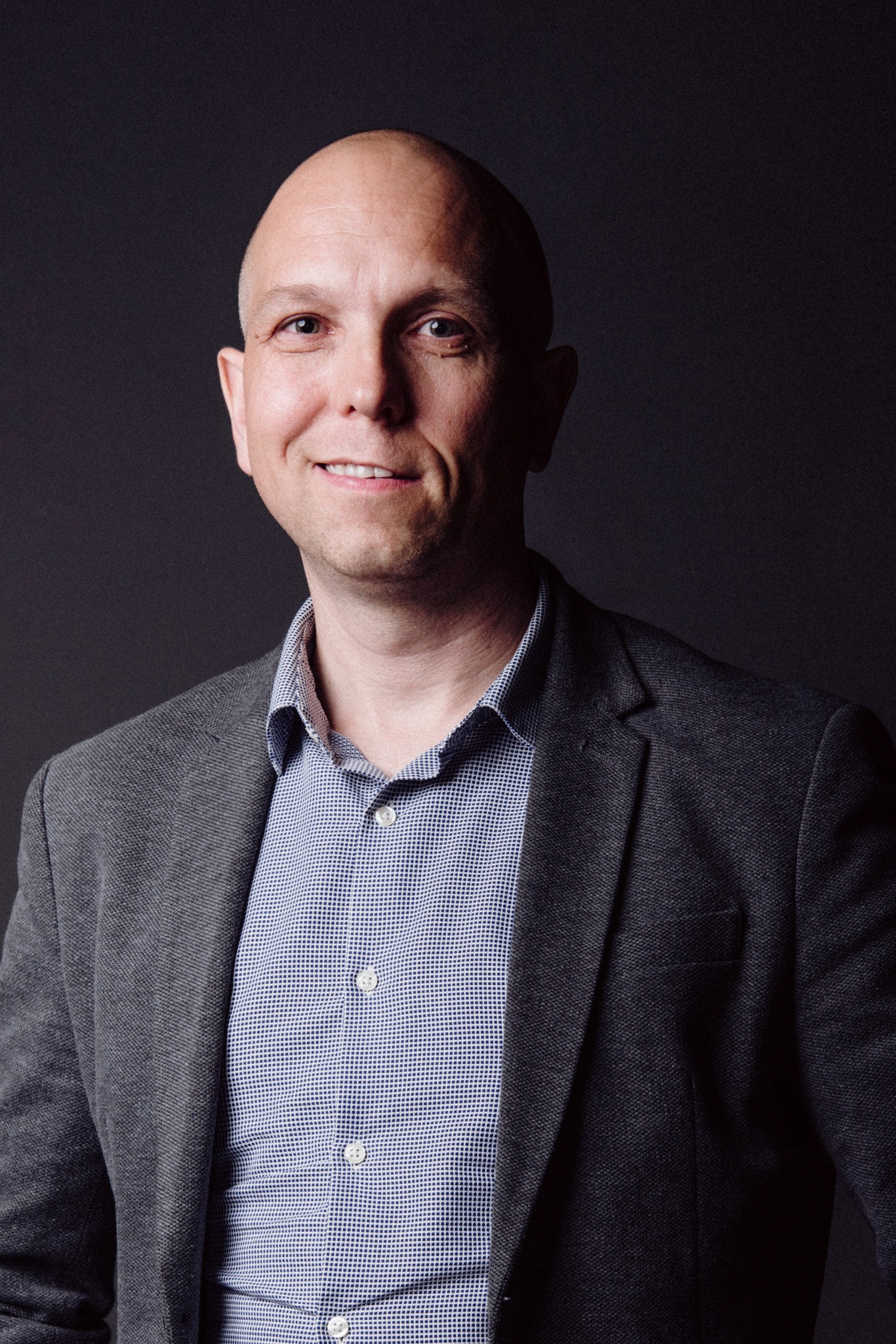 Portret uśmiechniętego mężczyzny w szarej marynarce i niebieskiej koszuli na ciemnoszarym tle.