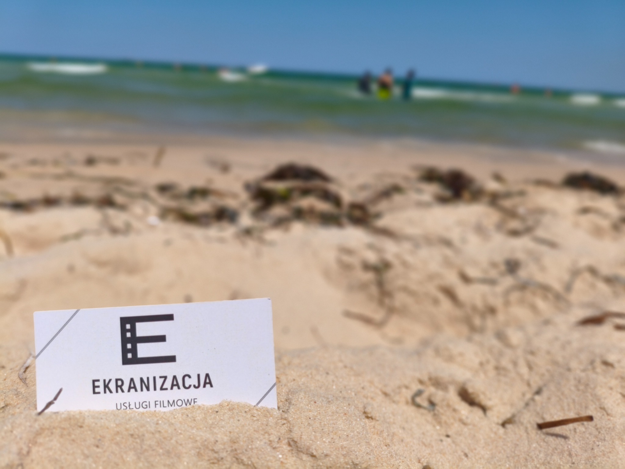 Wizytówka firmy 'Ekranizacja Usługi Filmowe' wbita w piasek na plaży. W tle morze z falami i sylwetki ludzi. Słoneczny dzień.