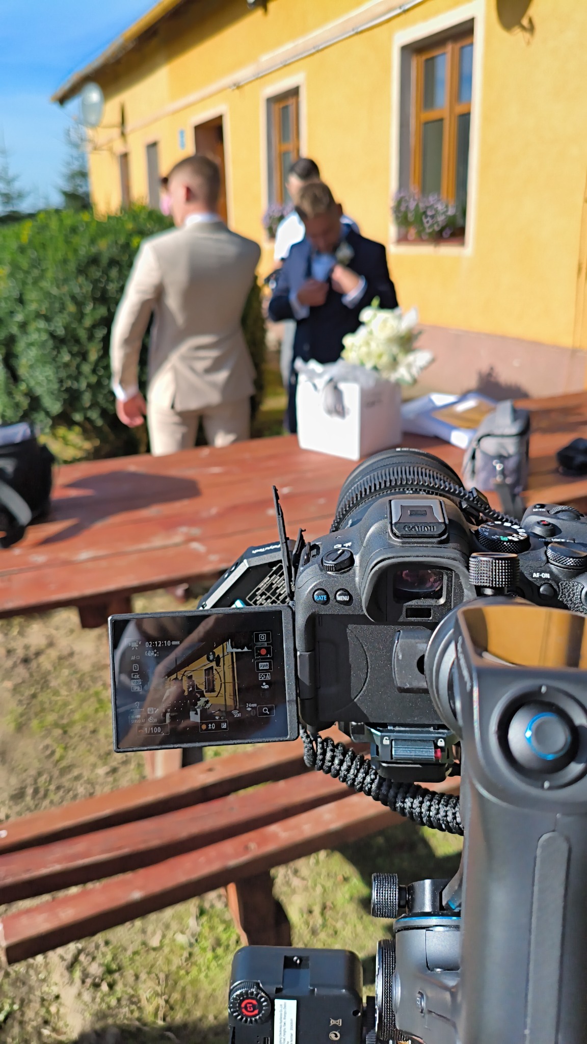 Profesjonalny aparat Canon na statywie, ekran LCD pokazuje ujęcie z przygotowań ślubnych. W tle para młoda i dekoracje na drewnianym stole. Scena zza kulis.