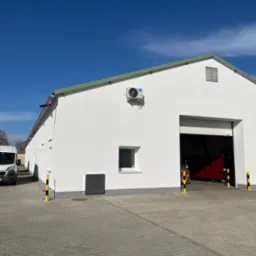 Biały budynek magazynowy z otwartą bramą, widoczny biały bus na kostce brukowej, błękitne niebo w tle.