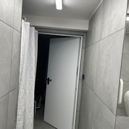 Firma OgólnoBudowlana Karol Szynkiewicz - Wykończona łazienka z szarymi płytkami, białą zasłoną prysznicową w romby i otwartymi białymi drzwiami.