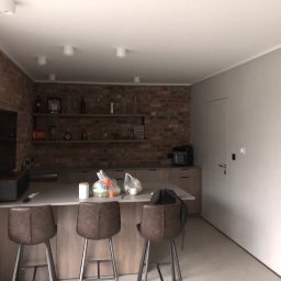 Firma OgólnoBudowlana Karol Szynkiewicz - Wnętrze kuchni z ceglaną ścianą, szarymi szafkami, wyspą z hokerami i świeżo pomalowaną szarą ścianą z ukrytymi drzwiami.