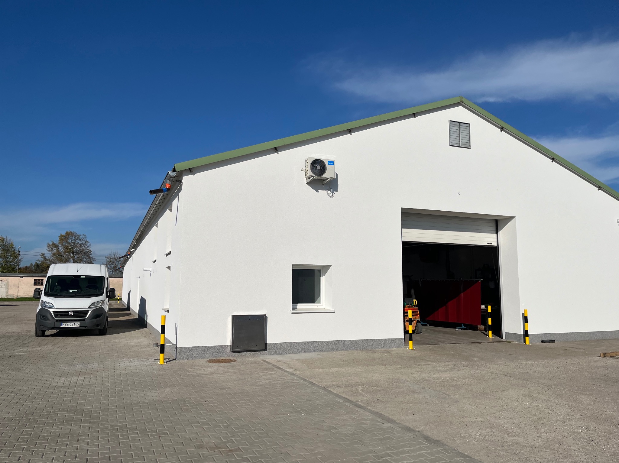 Biały budynek magazynowy z otwartą bramą, widoczny biały bus na kostce brukowej, błękitne niebo w tle.