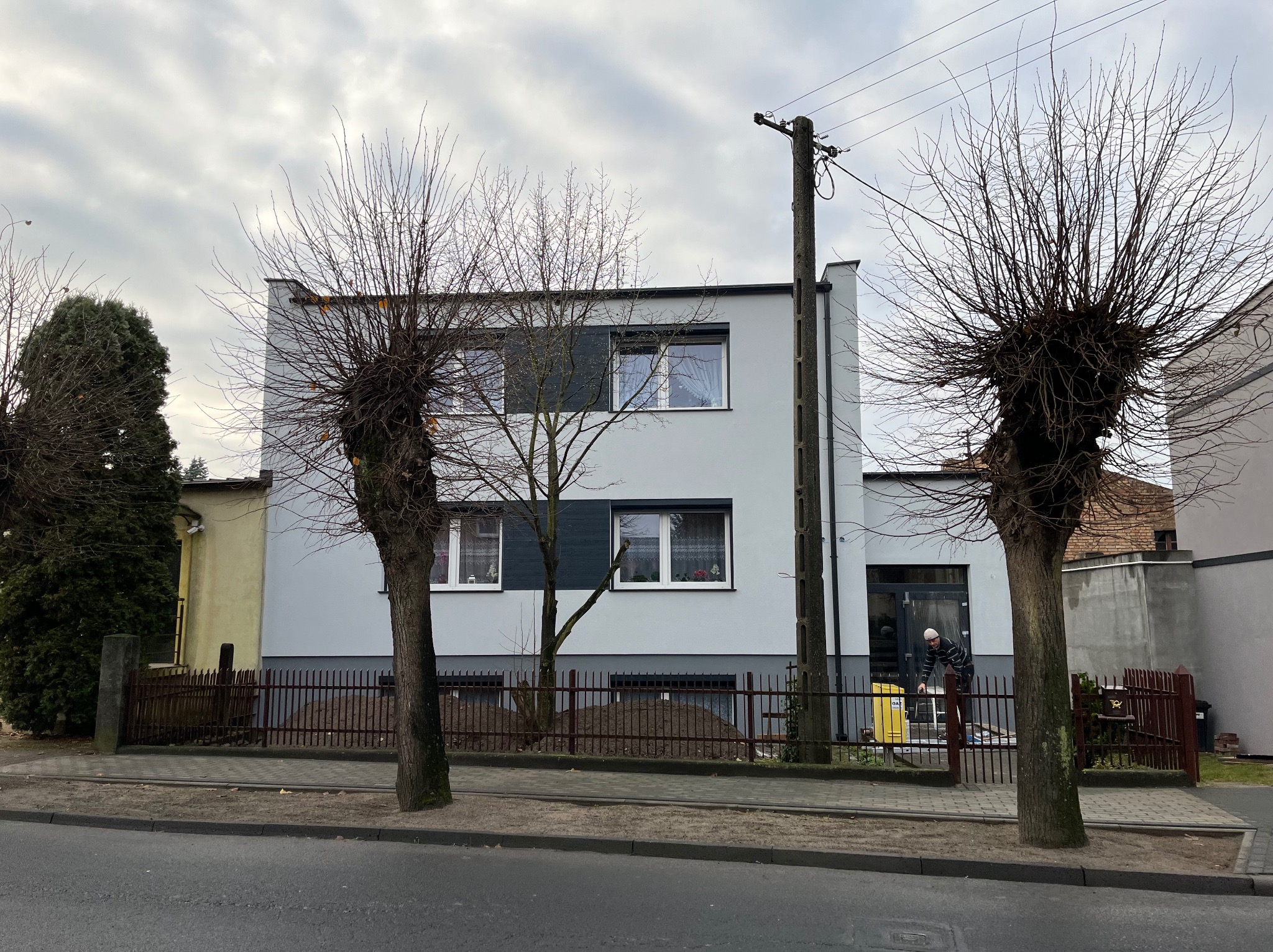 Pracownik w czapce i pasiastej bluzie przygotowuje sprzęt do pracy przed odnowioną, pomalowaną na szaro elewacją budynku, widoczne drzewa bez liści i słup elektryczny.