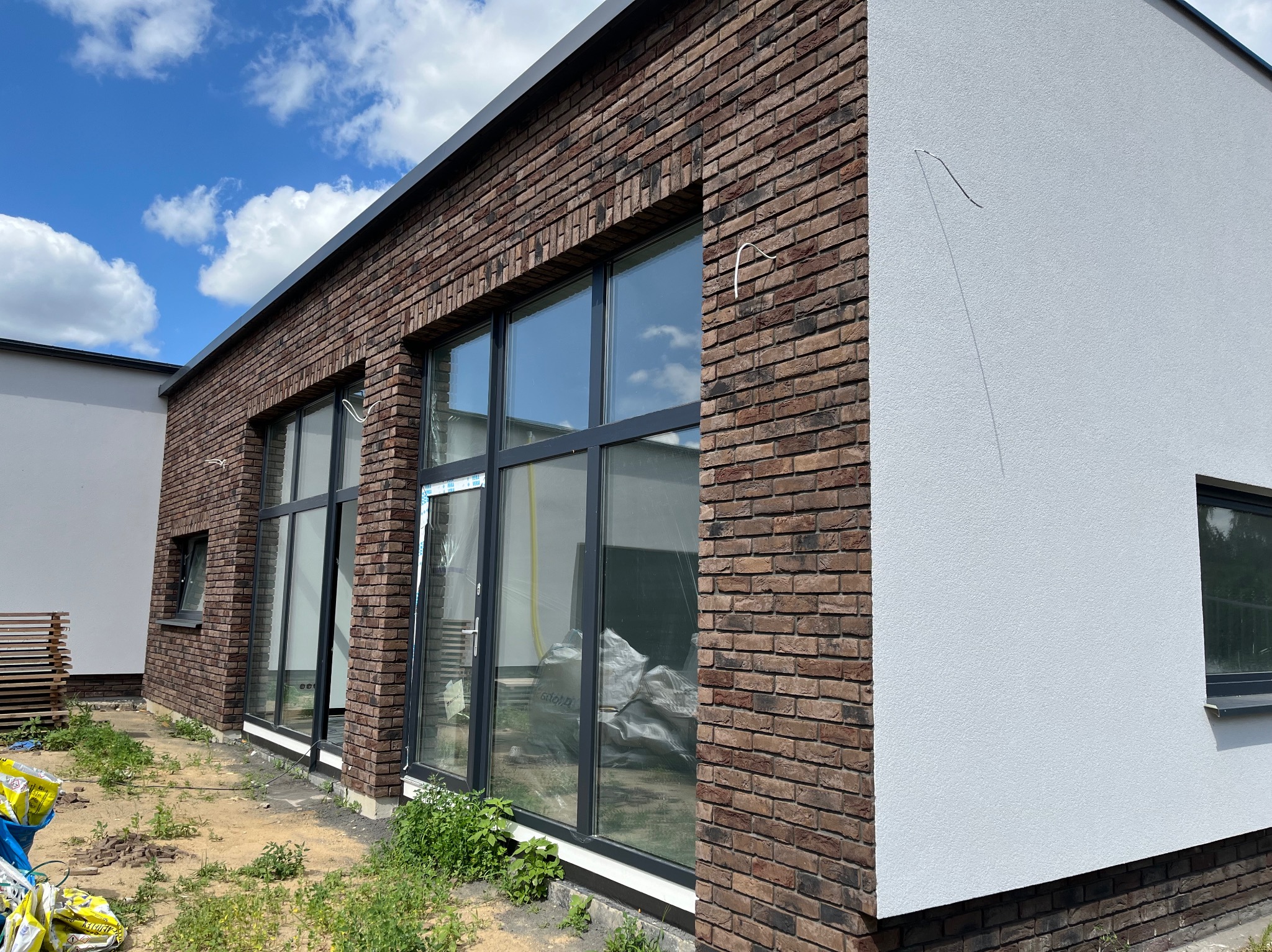 Nowoczesny dom z elewacją z ciemnej cegły i jasnym tynkiem mineralnym, duże okna z czarnymi ramami, widoczne worki z materiałami budowlanymi przed budynkiem.