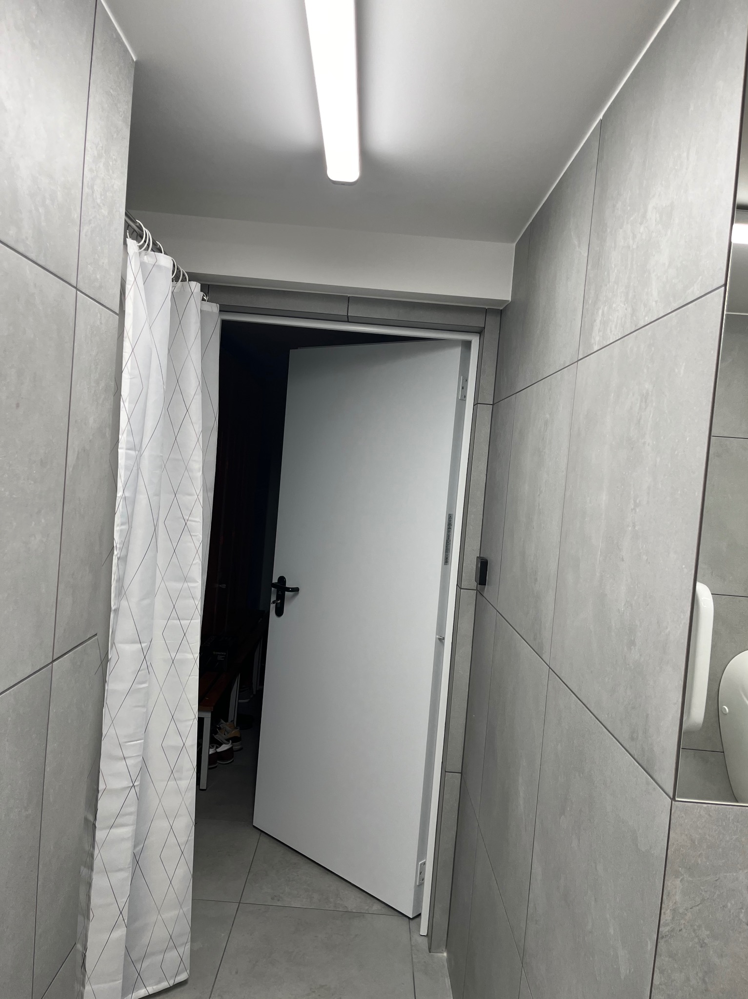 Wykończona łazienka z szarymi płytkami, białą zasłoną prysznicową w romby i otwartymi białymi drzwiami.