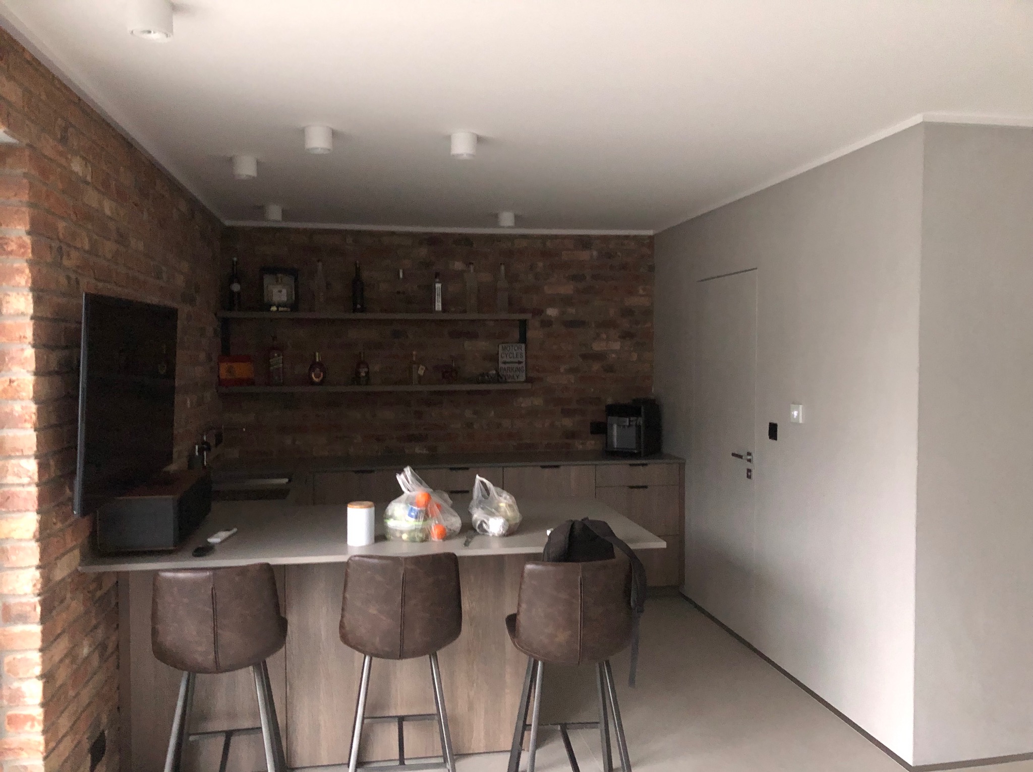 Wnętrze kuchni z ceglaną ścianą, szarymi szafkami, wyspą z hokerami i świeżo pomalowaną szarą ścianą z ukrytymi drzwiami.