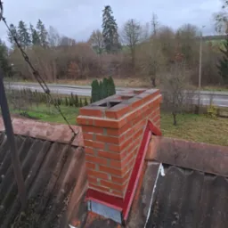 Ceglany komin z czerwoną metalową konstrukcją wsporczą na starym, bitumicznym dachu z widocznymi śladami napraw i uszczelnieniami, na tle pochmurnego nieba i drzew.