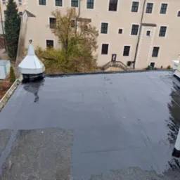 Widok z góry na płaski dach pokryty czarną hydroizolacją, z widocznym kominkiem wentylacyjnym i fragmentem starego pokrycia. W tle budynek z wieloma oknami i elementami architektonicznymi.