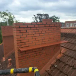 Murowany komin z czerwonej cegły na dachu pokrytym dachówką, widok z góry, fragment rusztowania z żółtymi pasami w kadrze.