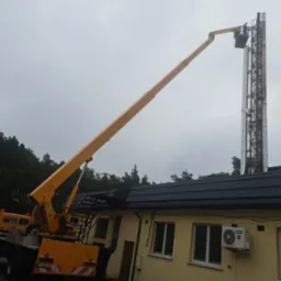 Pracownik na podnośniku koszowym przy wysokim kominie przemysłowym, widoczny budynek z klimatyzacją i las w tle.