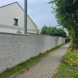 Długi, szary mur z bloczków betonowych, oddzielający posesję od chodnika z kostki brukowej, z fragmentem domu w tle i drzewem z pnączami przy chodniku.