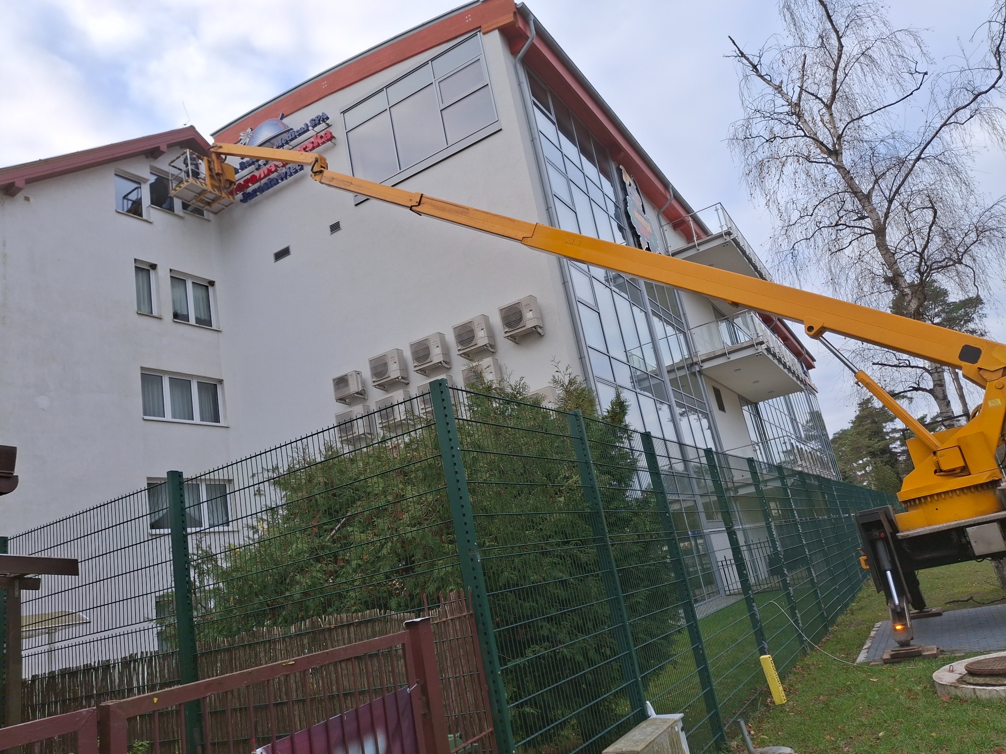 Montaż reklamy na elewacji budynku z użyciem podnośnika koszowego, widoczne klimatyzatory i ogrodzenie.