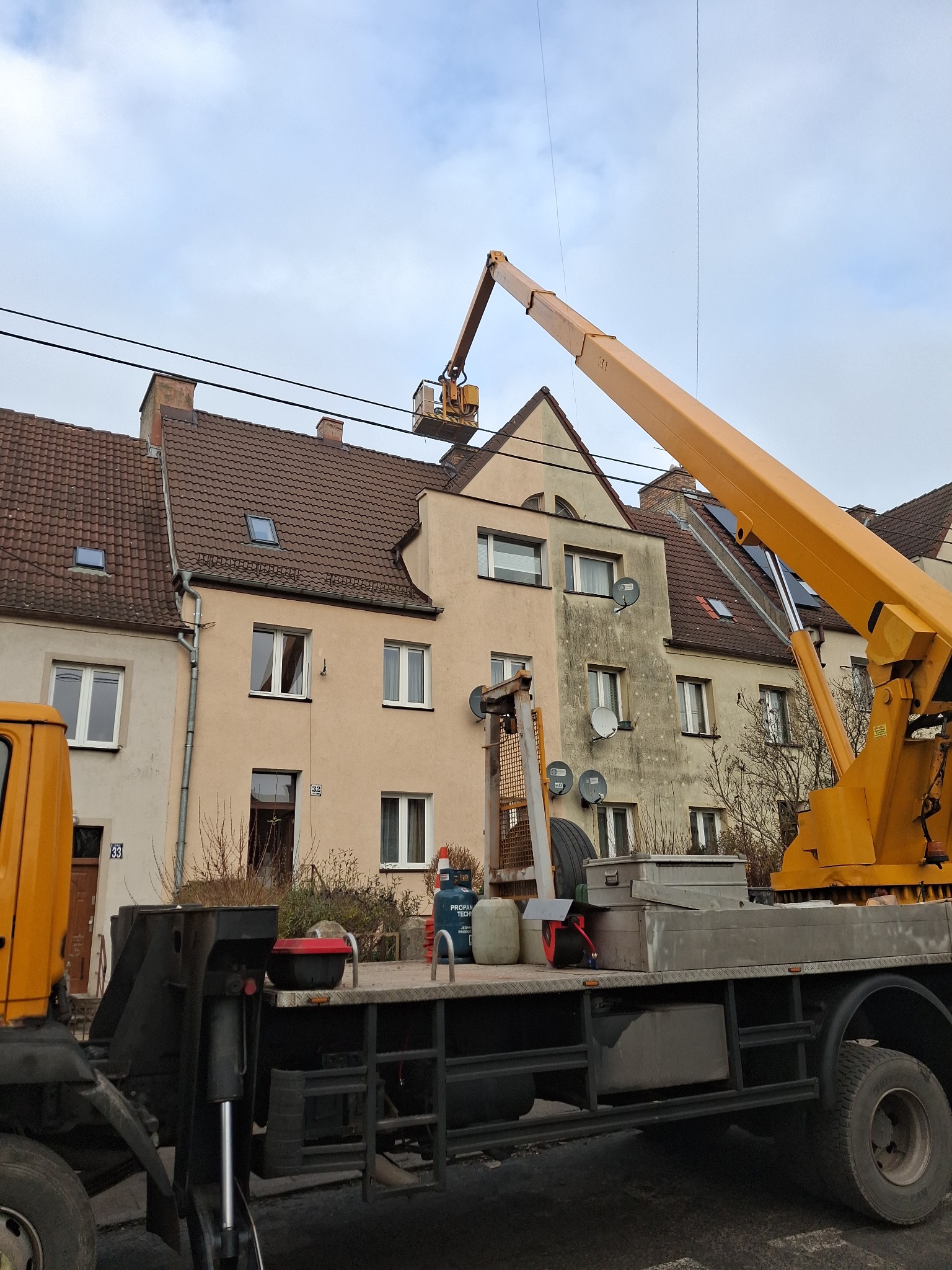 Pracownik w koszu żółtego podnośnika teleskopowego na tle dwuspadowego dachu budynku mieszkalnego z dachówki ceramicznej, widoczne okna dachowe i satelity na elewacji.