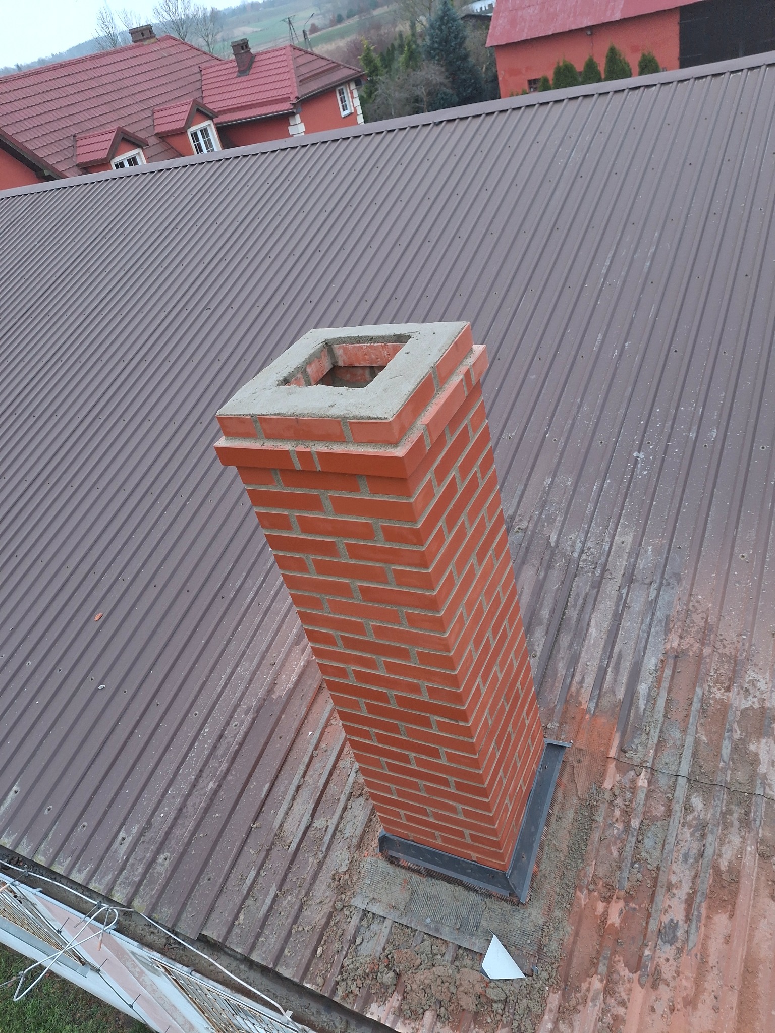 Nowy, murowany komin z cegły klinkierowej na brązowym, blaszanym dachu z widocznymi śladami montażu i pobliskimi budynkami mieszkalnymi.