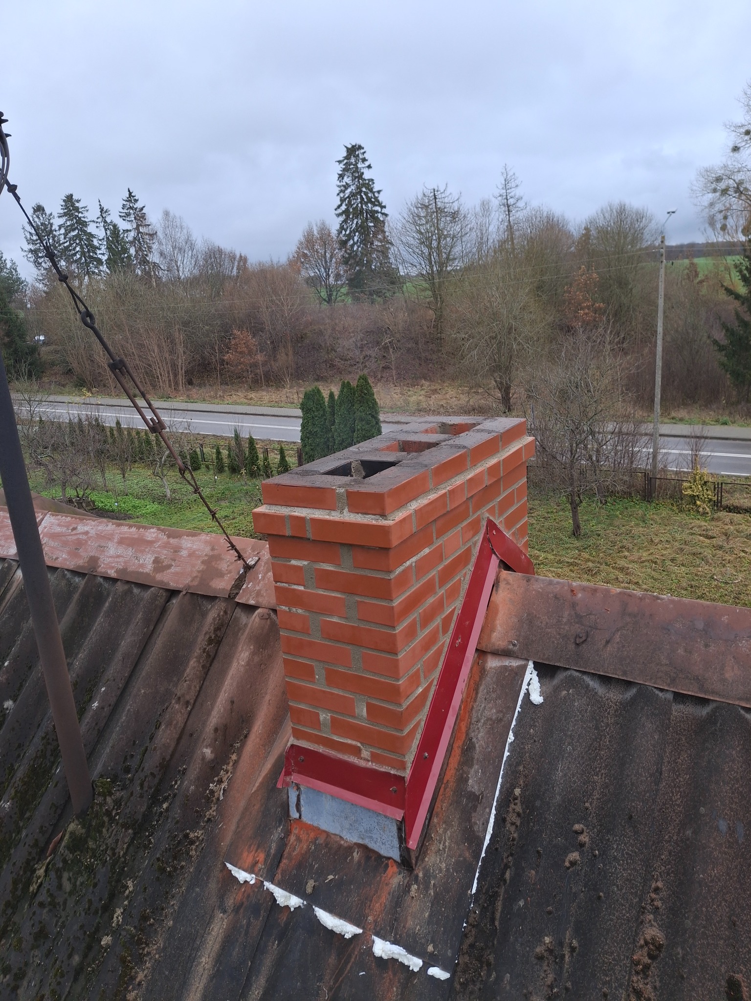 Ceglany komin z czerwoną metalową konstrukcją wsporczą na starym, bitumicznym dachu z widocznymi śladami napraw i uszczelnieniami, na tle pochmurnego nieba i drzew.