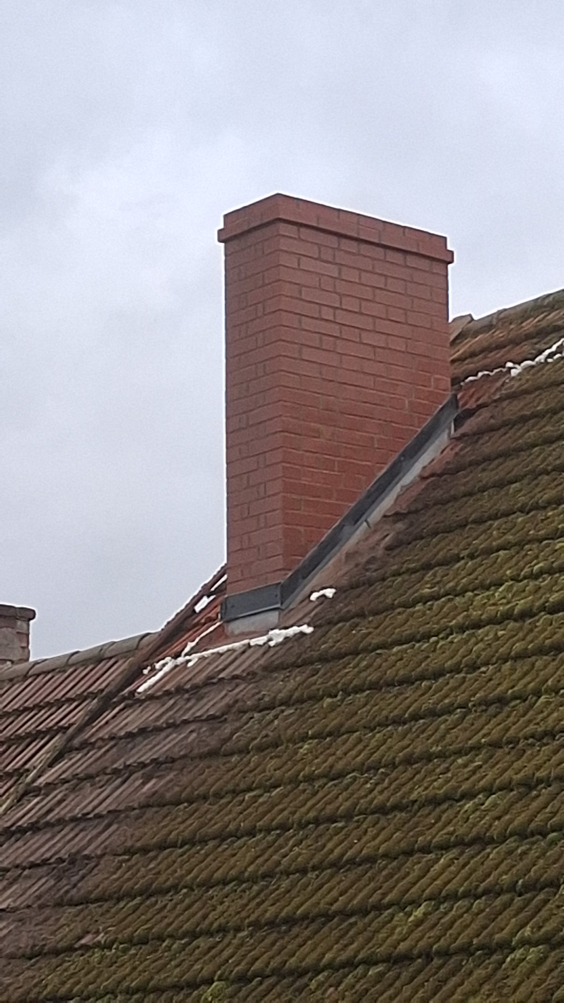 Ceglany komin wystający z dachu pokrytego dachówką częściowo porośniętą mchem, z widocznymi fragmentami śniegu.