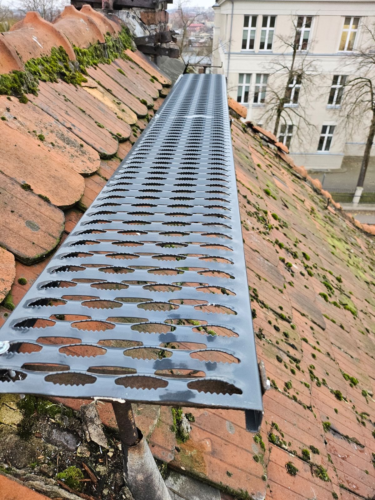 Szara, perforowana metalowa kładka na starym, ceglanym dachu pokrytym mchem, widok z góry, w tle budynek z oknami.