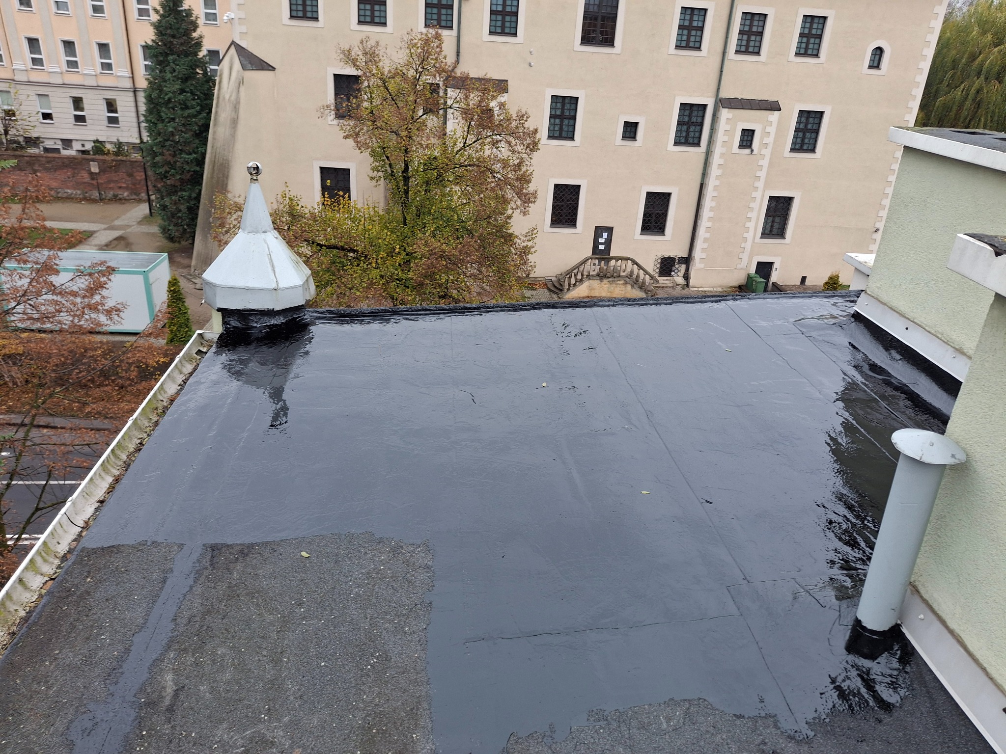 Widok z góry na płaski dach pokryty czarną hydroizolacją, z widocznym kominkiem wentylacyjnym i fragmentem starego pokrycia. W tle budynek z wieloma oknami i elementami architektonicznymi.