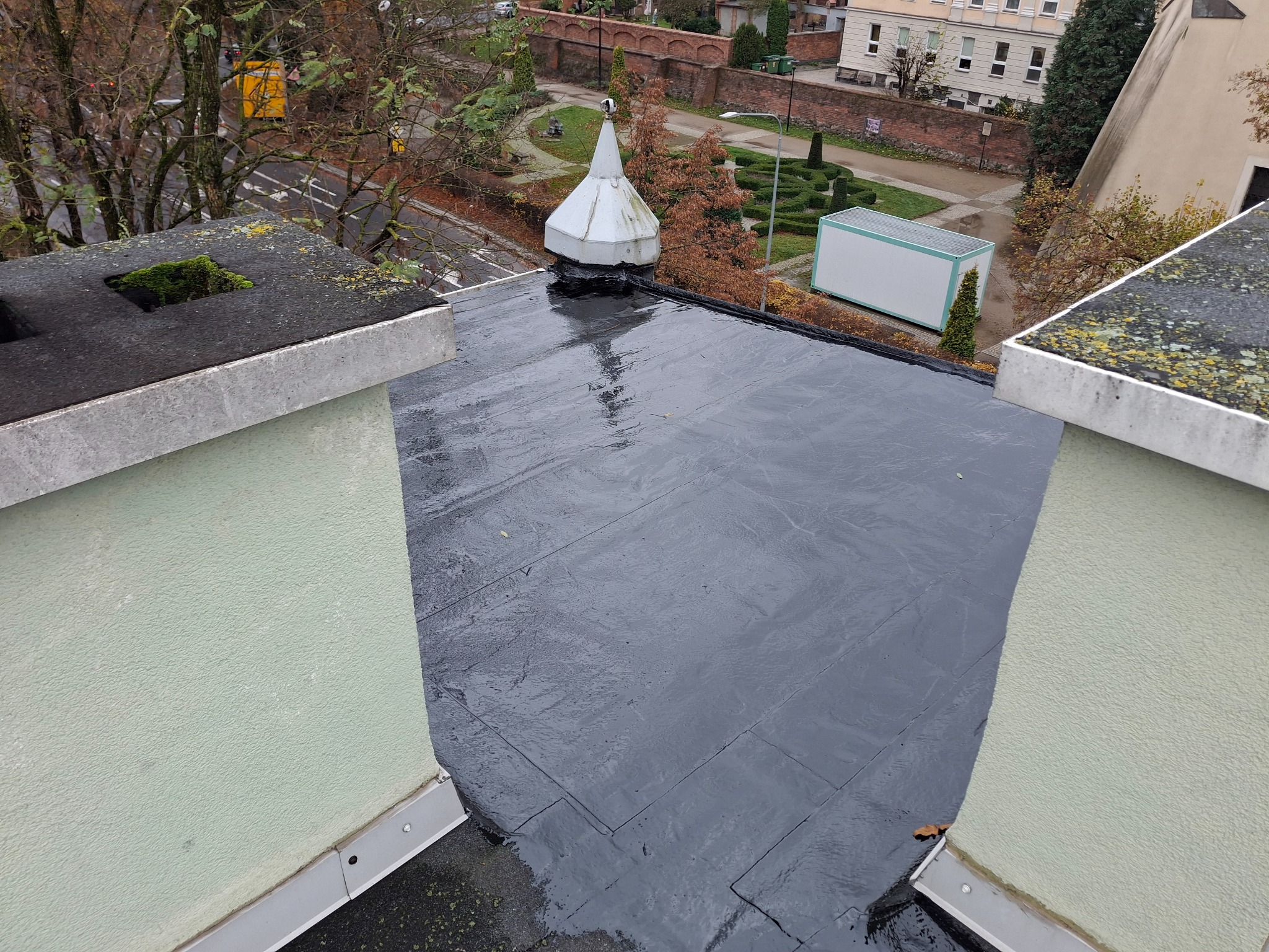 Czarny, płaski dach pokryty papą bitumiczną po deszczu, widok z góry na detale wykończenia attyki i otoczenie budynku.