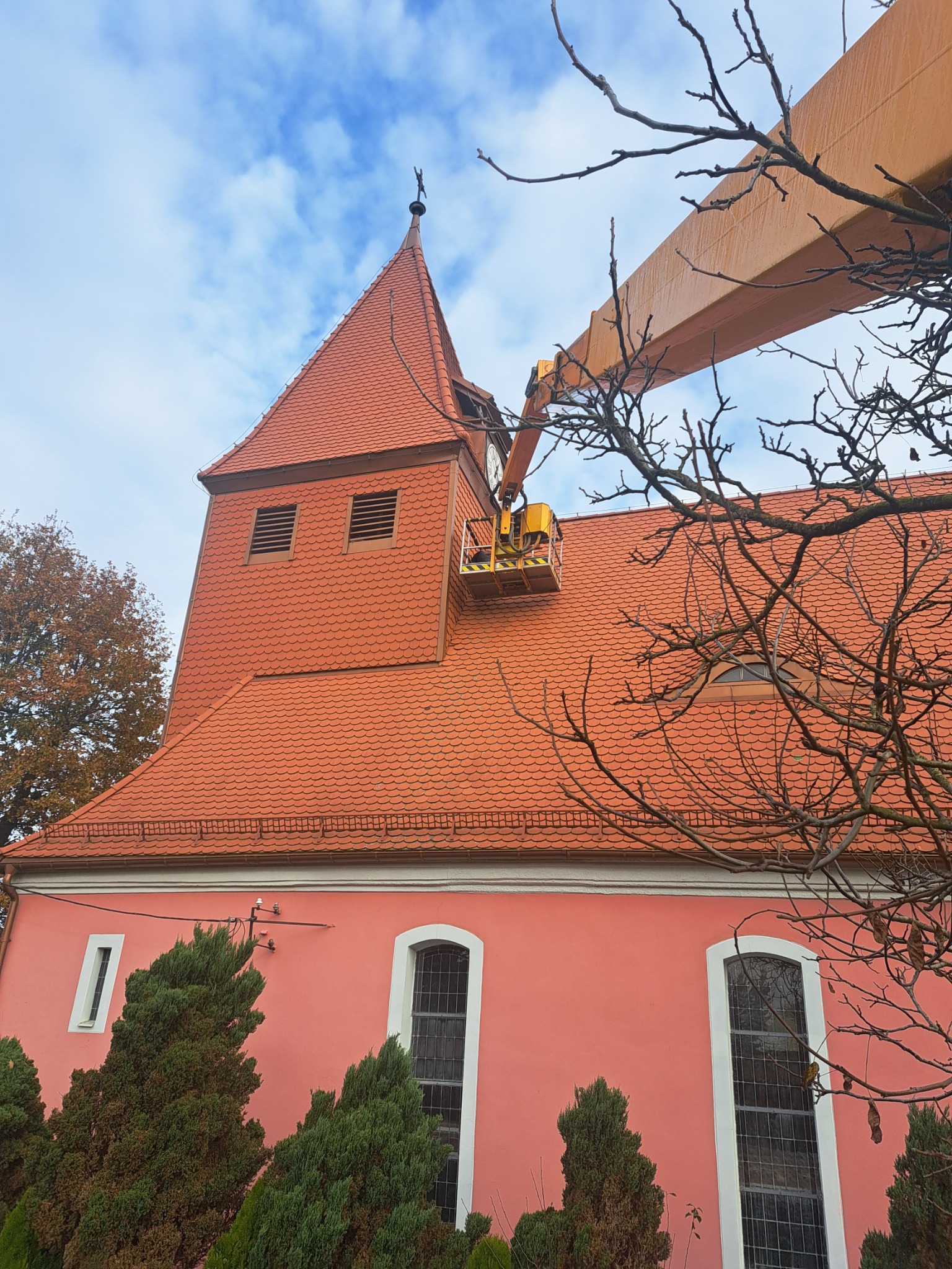 Prace na wysokościach przy kościele z użyciem podnośnika koszowego, widoczne elementy elewacji, dachu z dachówki ceramicznej i iglica na wieży, drzewa w tle.