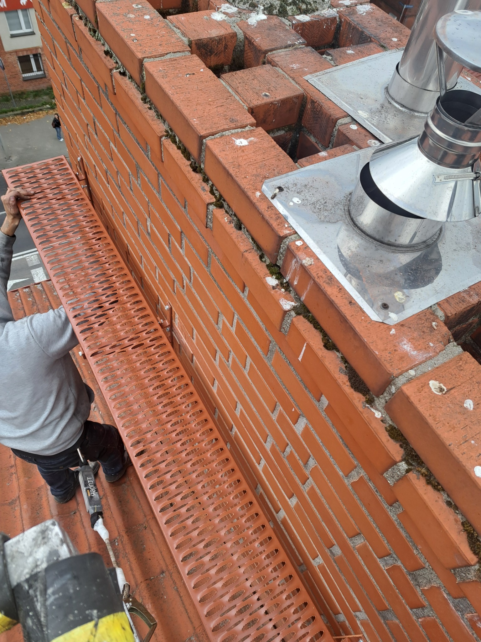 Murarz montuje pomost roboczy z perforowanej blachy przy kominie z czerwonej cegły, widoczne stalowe obróbki kominowe i fragment dachu.