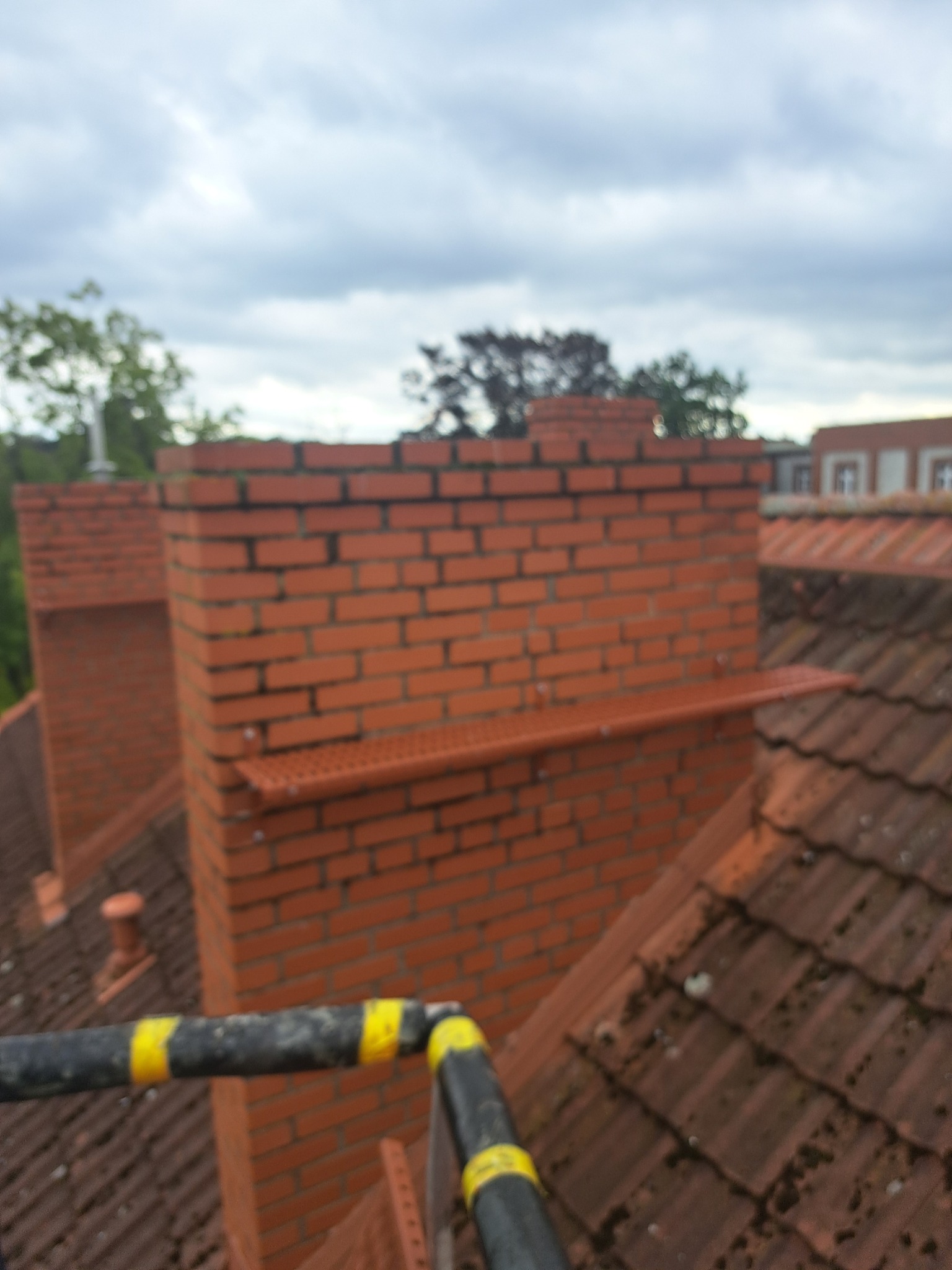 Murowany komin z czerwonej cegły na dachu pokrytym dachówką, widok z góry, fragment rusztowania z żółtymi pasami w kadrze.
