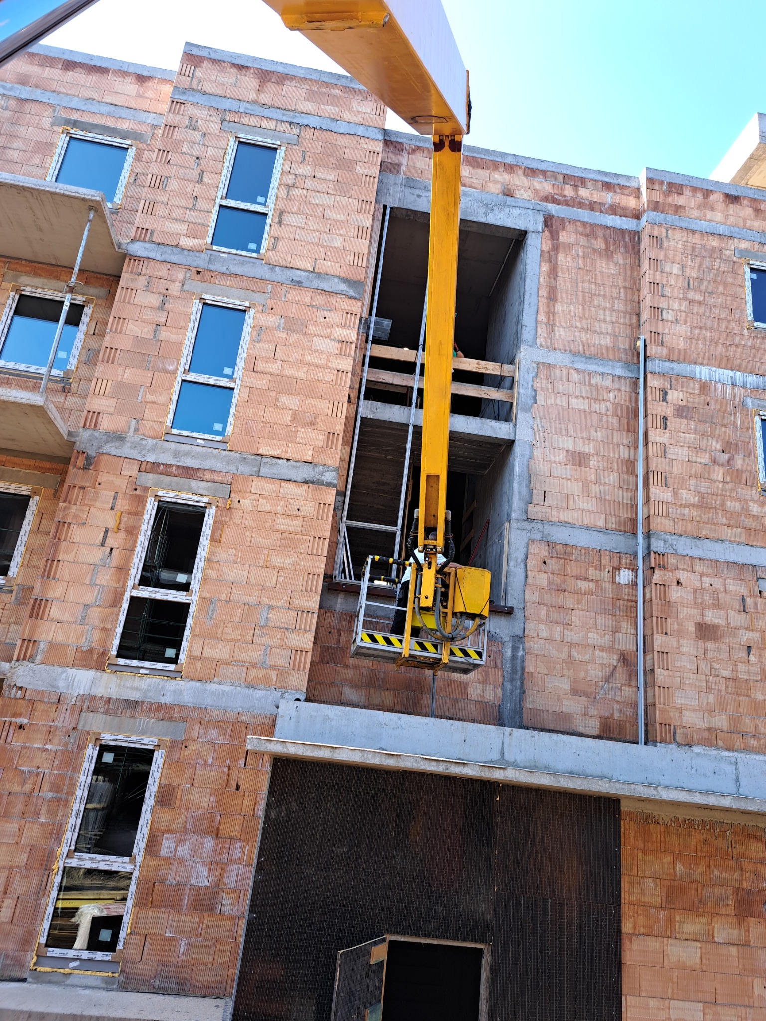 Budowa wielopiętrowego budynku z czerwonej cegły, widoczny podnośnik koszowy z pracownikiem na wysokości, okna w trakcie montażu, surowy stan.
