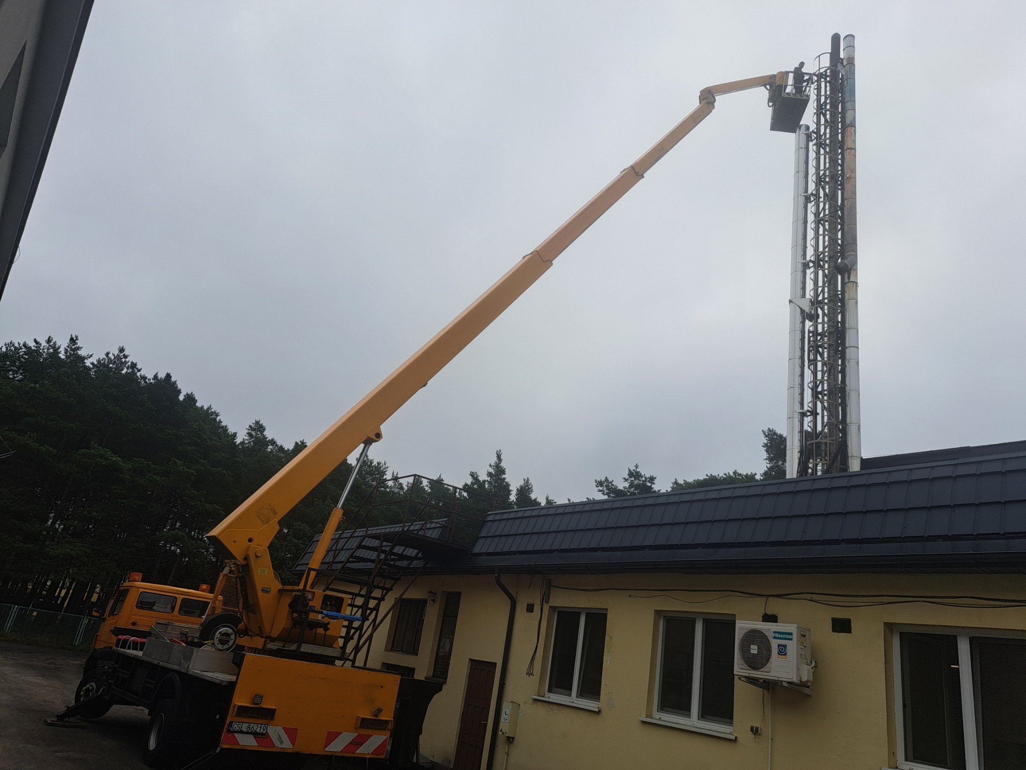Pracownik na podnośniku koszowym przy wysokim kominie przemysłowym, widoczny budynek z klimatyzacją i las w tle.