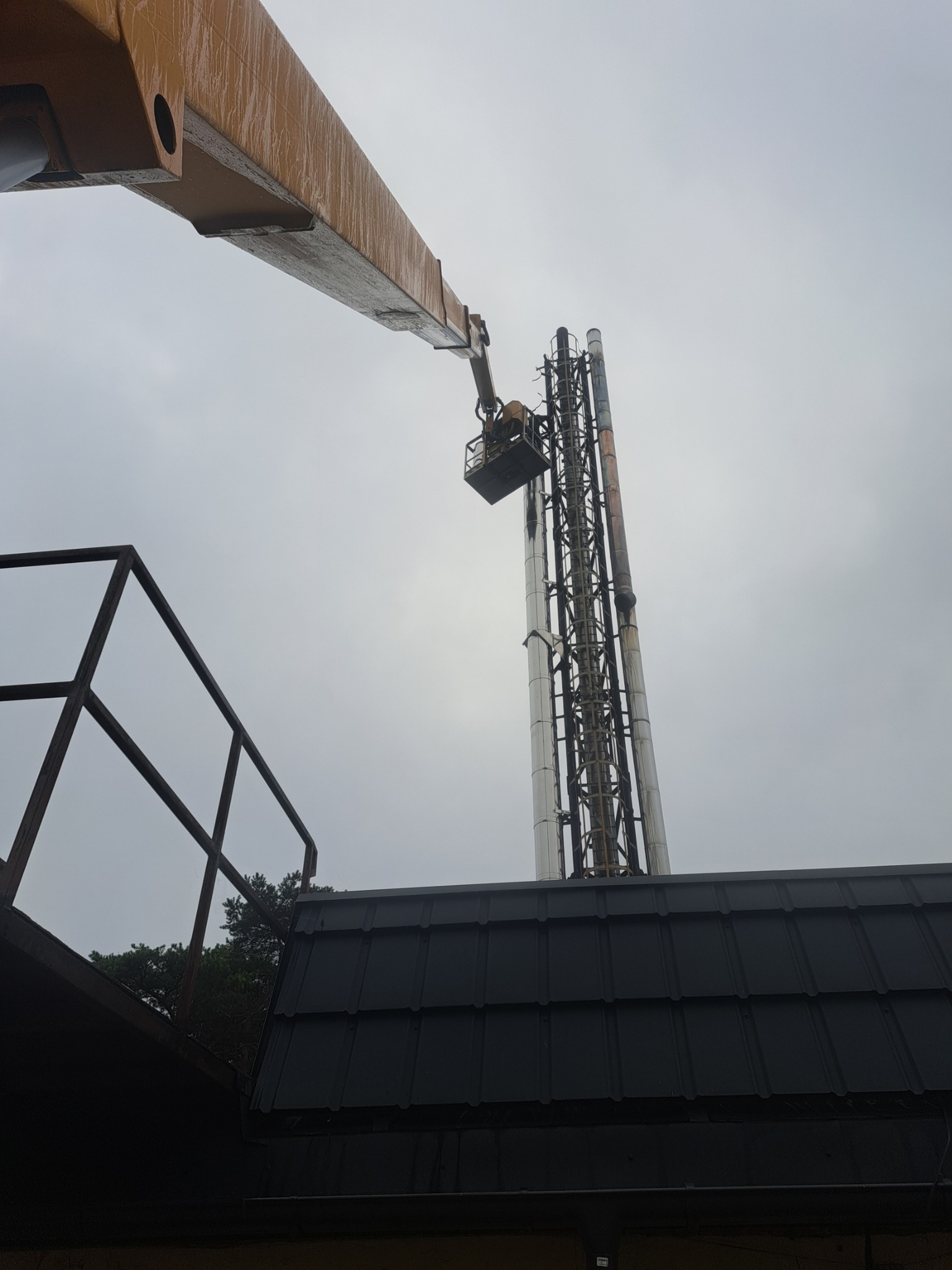 Montaż lub naprawa wysokiego komina przemysłowego z platformy podnośnika koszowego, widok z dołu na tle pochmurnego nieba.