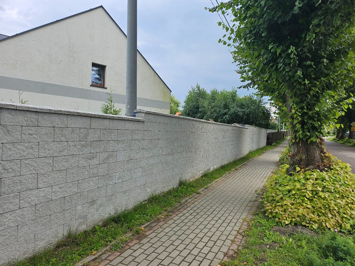 Długi, szary mur z bloczków betonowych, oddzielający posesję od chodnika z kostki brukowej, z fragmentem domu w tle i drzewem z pnączami przy chodniku.