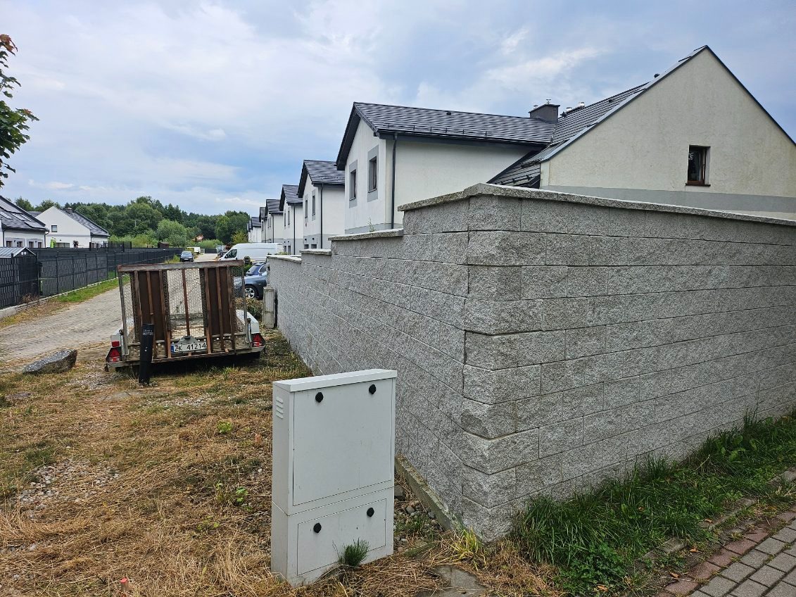 Szary, kamienny mur oporowy oddzielający posesje z nowymi domami jednorodzinnymi w tle, widoczna skrzynka elektryczna i przyczepka samochodowa.