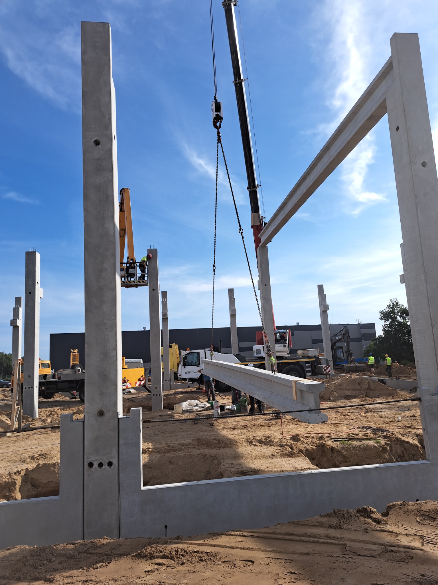 Montaż prefabrykowanych, żelbetowych słupów i belek konstrukcyjnych na placu budowy z użyciem dźwigu, widoczne przygotowane fundamenty, w tle maszyny budowlane i pracownicy w odblaskowych kamizelkach.