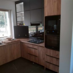 KU-GA Meble na wymiar - Nowoczesna kuchnia z drewnianymi frontami szafek, czarnymi elementami i blatem z płyty drewnopodobnej, widok na okno z żaluzjami.