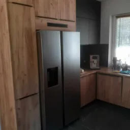 Nowoczesna kuchnia z drewnianymi frontami szafek, czarnymi elementami i dużą, dwudrzwiową lodówką ze stali nierdzewnej.