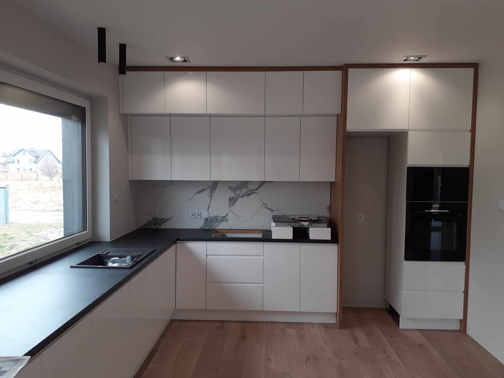 Nowoczesna kuchnia z białymi szafkami, czarnym blatem i marmurowym backsplashiem, oświetlona wpuszczanymi lampami i z widokiem na zewnątrz przez duże okno.