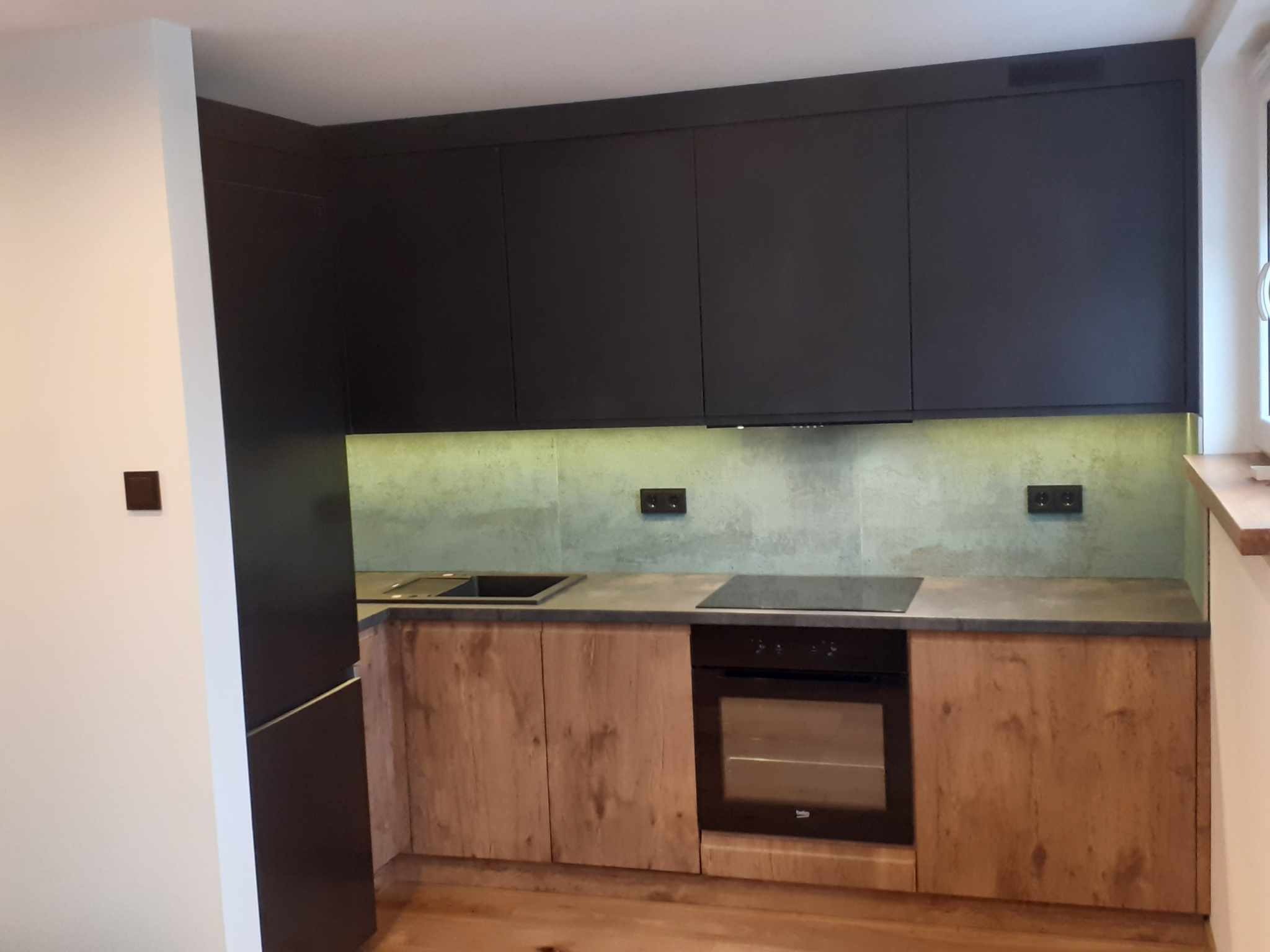 Nowoczesna kuchnia z ciemnymi, wiszącymi szafkami i drewnopodobnymi frontami dolnych szafek, podświetlona listwą LED pod szafkami górnymi, z ciemnym blatem i płytą indukcyjną oraz piekarnikiem...