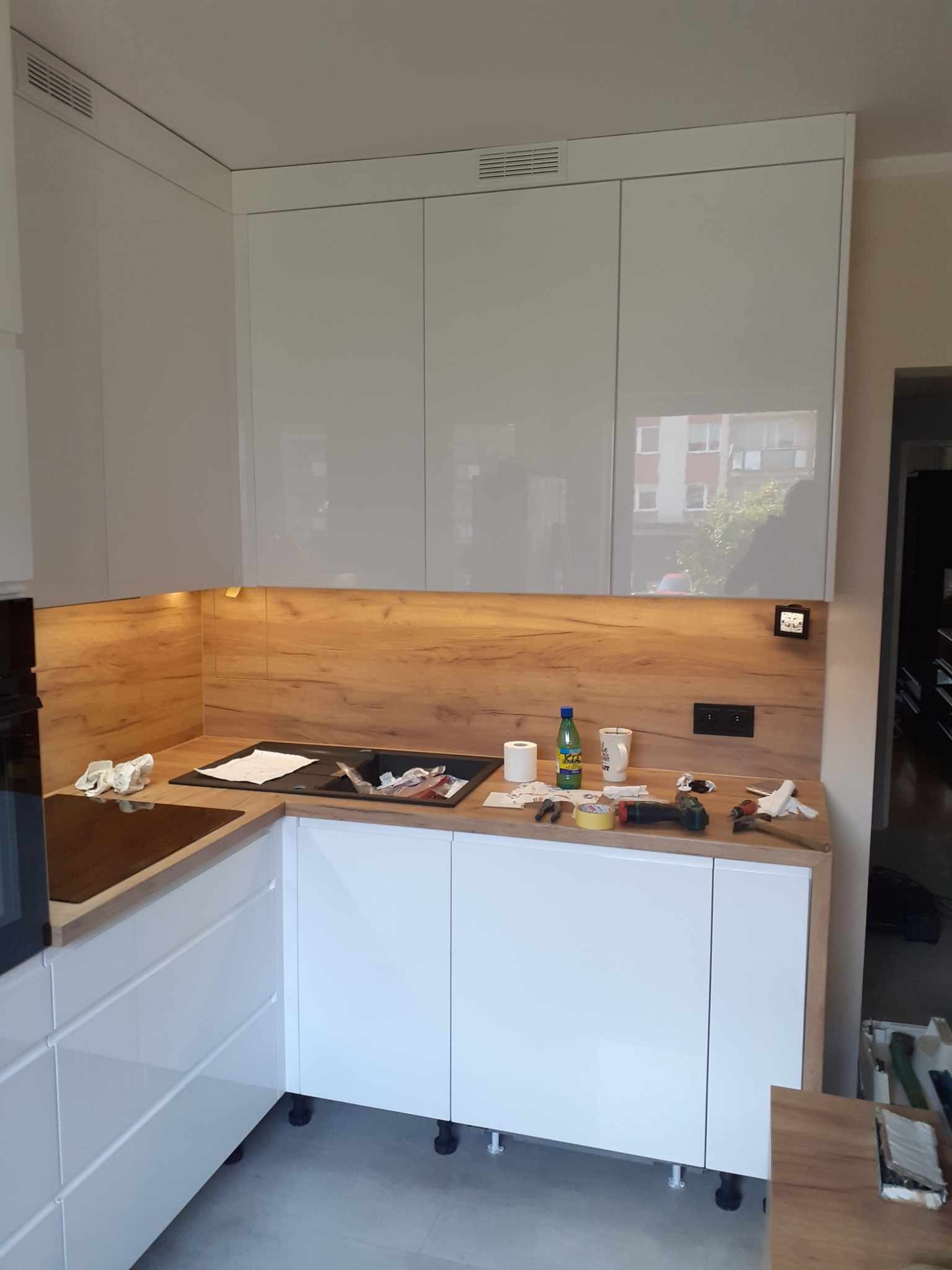 Montaż białej, lakierowanej kuchni z drewnianym blatem i panelem ściennym; widoczne narzędzia i akcesoria montażowe na blacie.