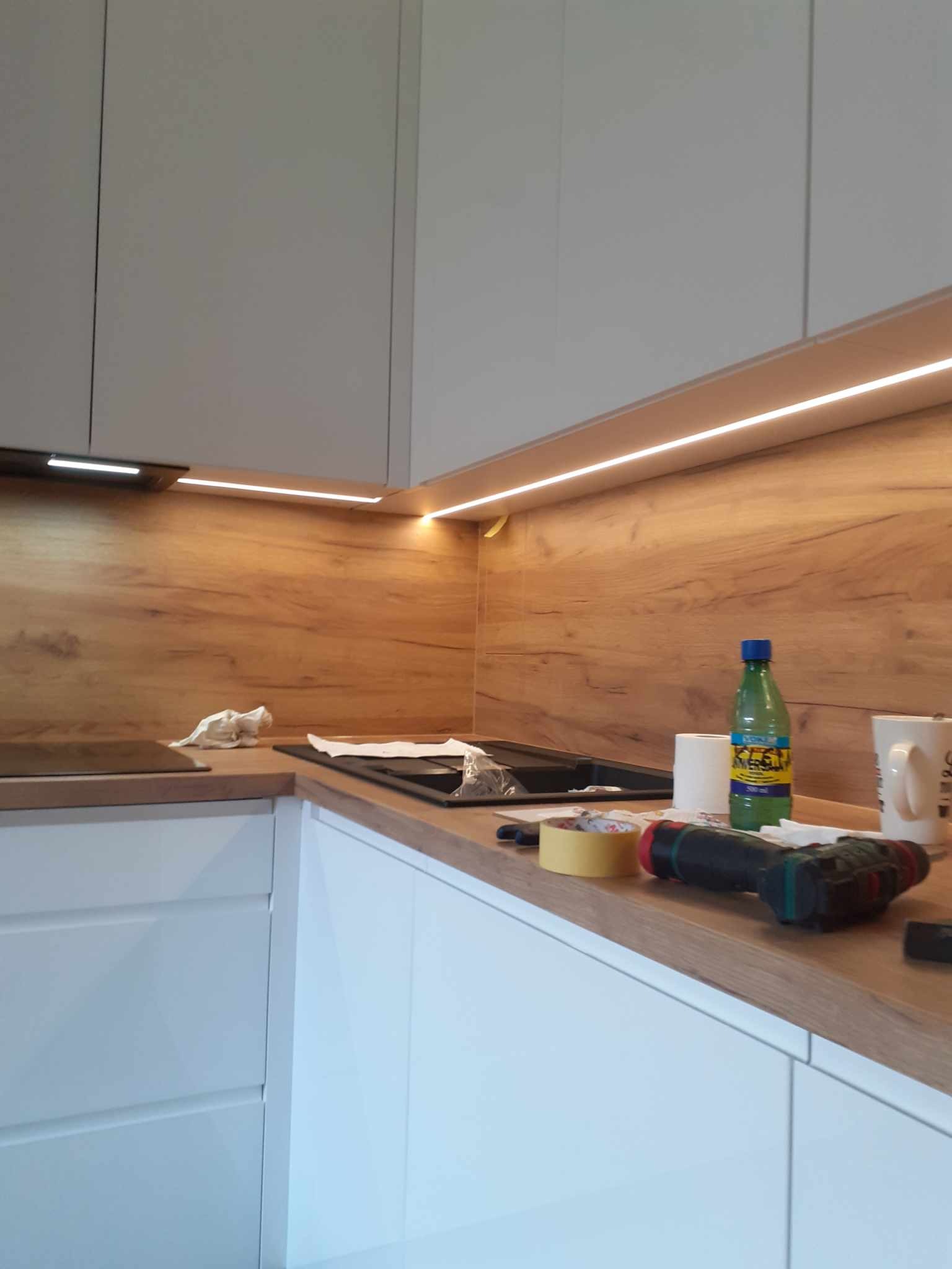 Montaż oświetlenia LED pod szafkami wiszącymi w kuchni z drewnianym blatem i okładziną ścienną, widoczne narzędzia i akcesoria budowlane.