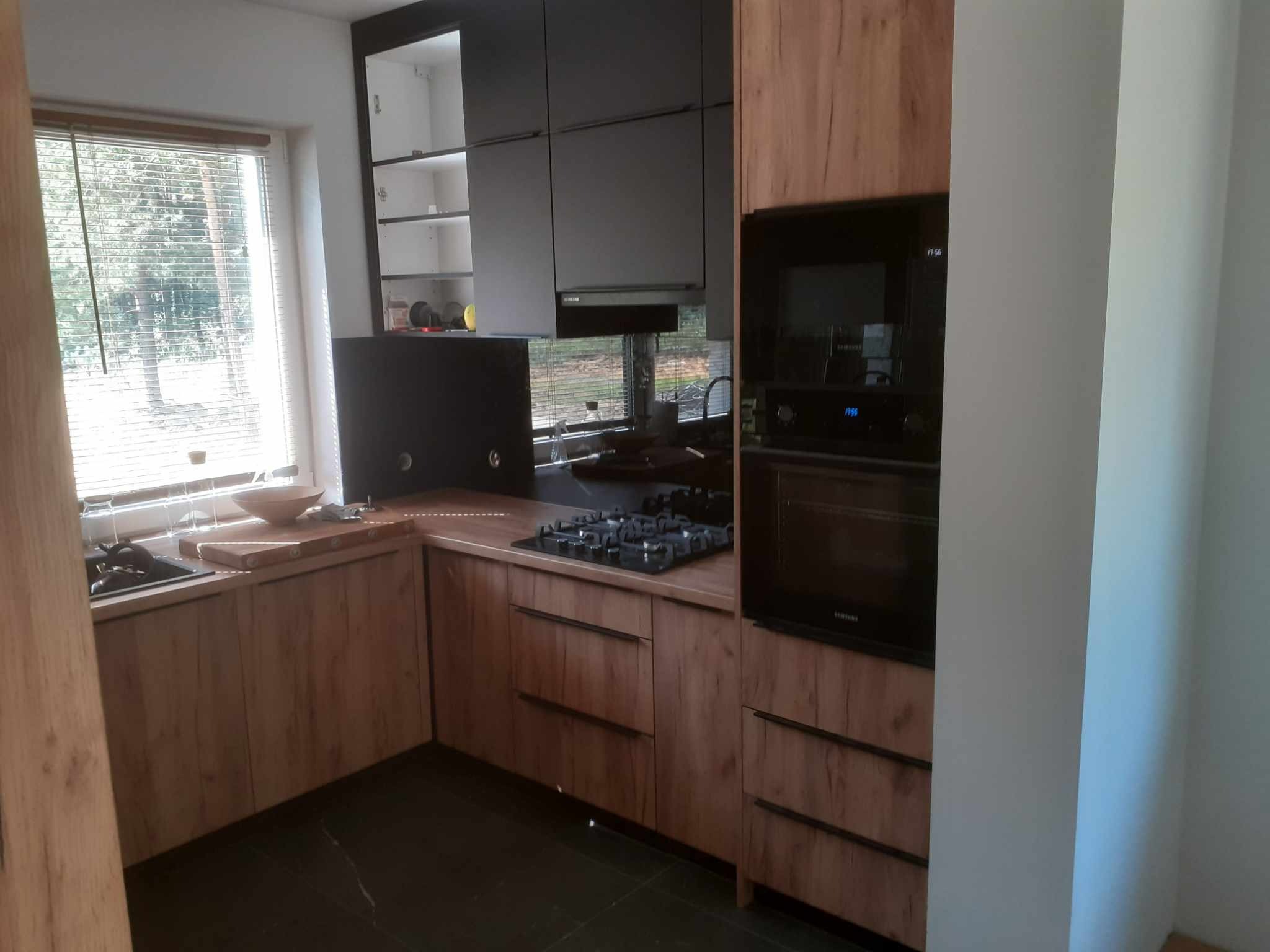 Nowoczesna kuchnia z drewnianymi frontami szafek, czarnymi elementami i blatem z płyty drewnopodobnej, widok na okno z żaluzjami.