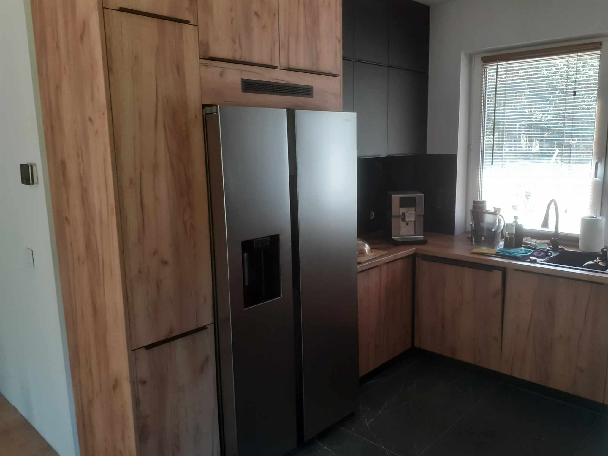 Nowoczesna kuchnia z drewnianymi frontami szafek, czarnymi elementami i dużą, dwudrzwiową lodówką ze stali nierdzewnej.