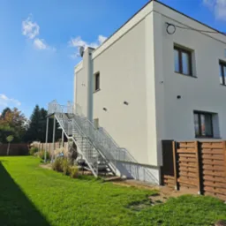 Nowoczesny, dwukondygnacyjny budynek z białym tynkiem i zewnętrznymi metalowymi schodami ewakuacyjnymi, widocznymi z boku, na tle zielonego trawnika i błękitnego nieba z lekkimi chmurami.