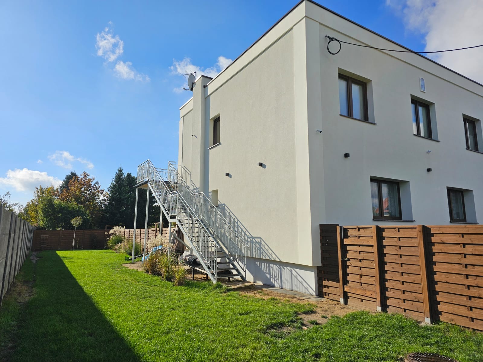 Nowoczesny, dwukondygnacyjny budynek z białym tynkiem i zewnętrznymi metalowymi schodami ewakuacyjnymi, widocznymi z boku, na tle zielonego trawnika i błękitnego nieba z lekkimi chmurami.