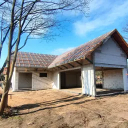 Surowy stan budynku z białego bloczka z drewnianą konstrukcją dachu i częściowo ułożoną dach&oacute;wką, widoczne drzewa bez liści i błękitne niebo.