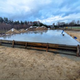 DarBet - Świeżo wylany fundament z drewnianym szalunkiem, widoczne hałdy piasku z resztkami śniegu i pochmurne niebo w tle.