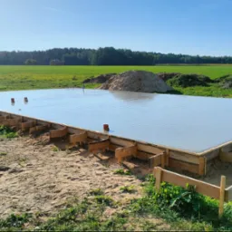 Świeżo wylany, gładki fundament betonowy z drewnianym szalunkiem, wystającymi rurami instalacyjnymi, na tle zielonego pola i lasu.