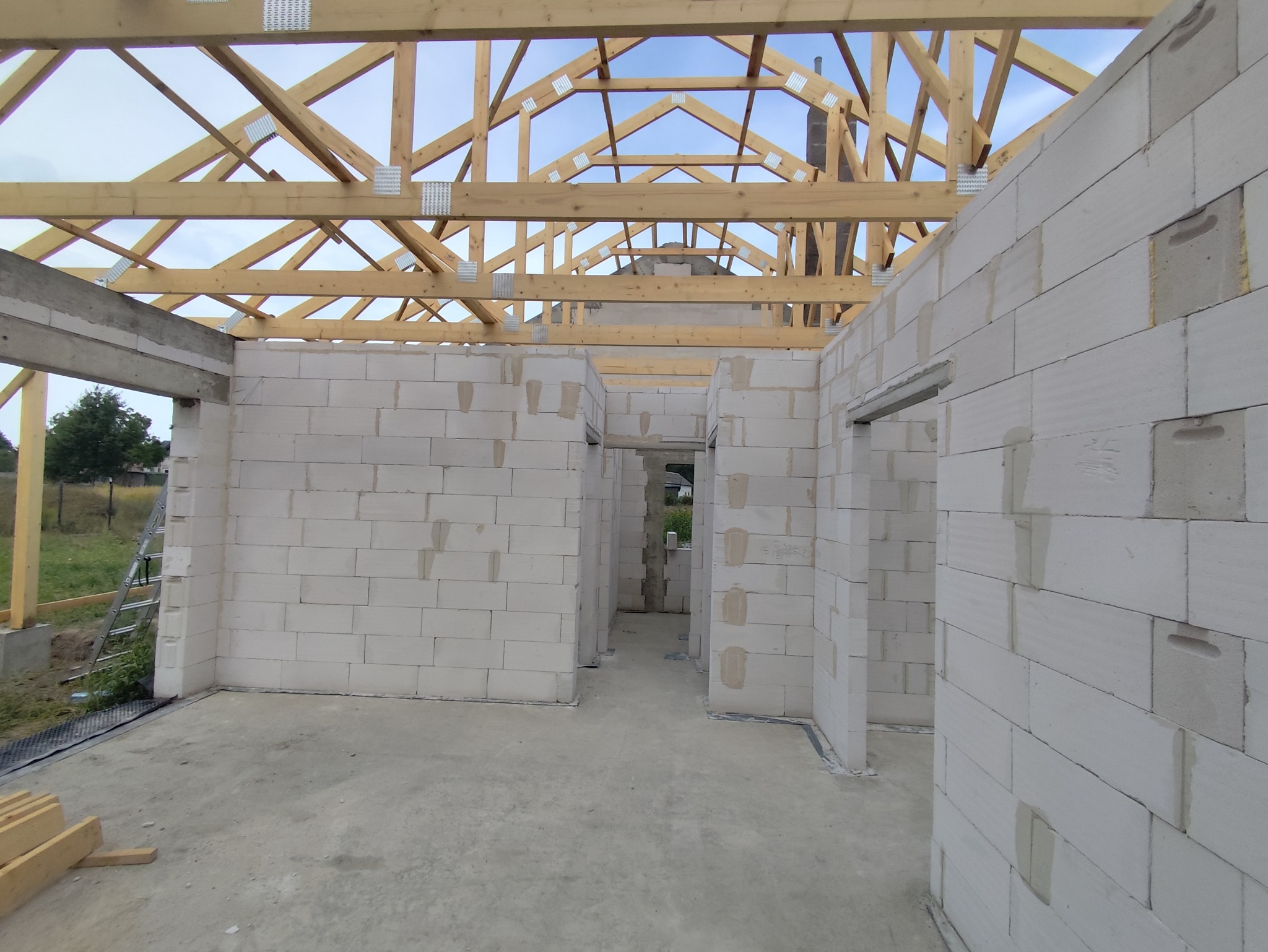 Wnętrze surowego domu w trakcie budowy: ściany z białych bloczków, widoczna drewniana konstrukcja dachu z metalowymi łącznikami, surowy betonowy podkład, widok na zewnątrz przez otwory okienne.
