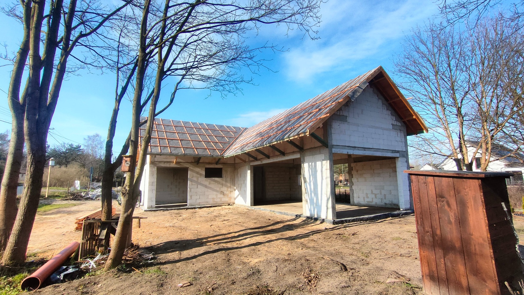 Surowy stan budynku z białego bloczka z drewnianą konstrukcją dachu i częściowo ułożoną dachówką, widoczne drzewa bez liści i błękitne niebo.