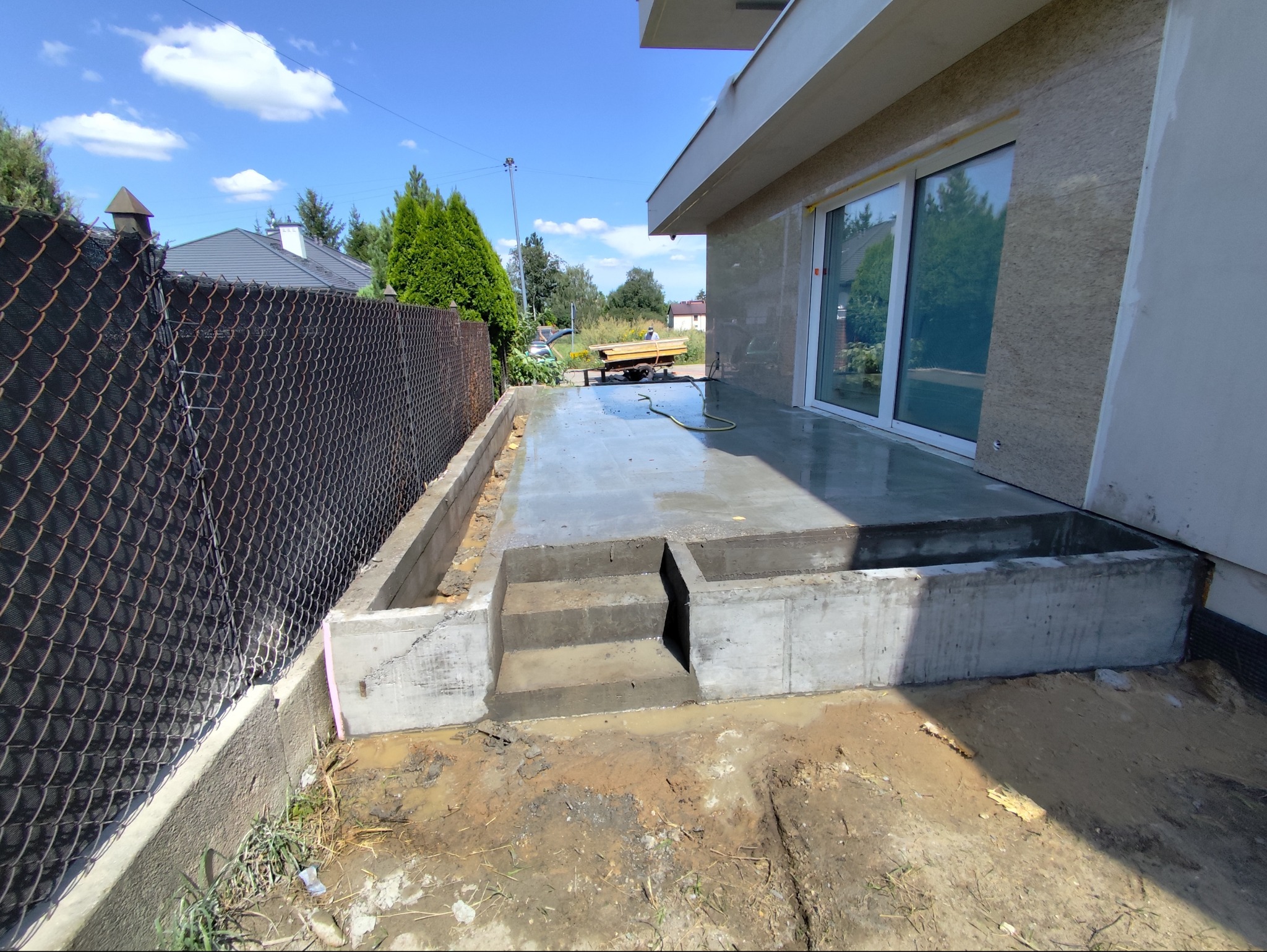 Świeżo wylany, mokry betonowy taras z trzema stopniami, widok z boku, z widoczną siatką ogrodzeniową i przygotowanym podłożem.